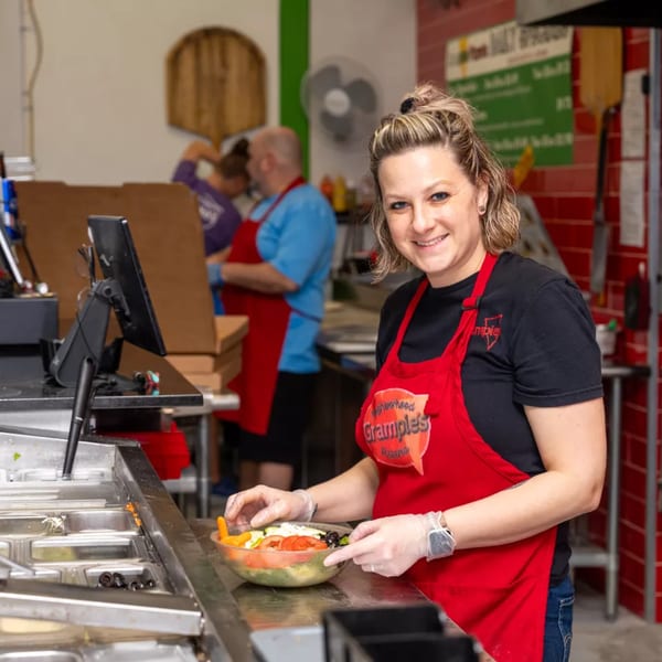 Smiling Grampie's Pizzeria employee in a red apron preparing a fresh salad behind the counter.