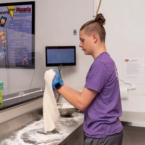 A Grampie's Pizzeria employee in a purple shirt and blue gloves stretches pizza dough over a floured counter.