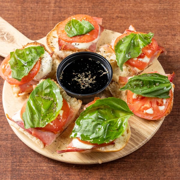 Six bruschetta pieces with tomato, mozzarella, prosciutto, and basil, surrounding a balsamic glaze dip.