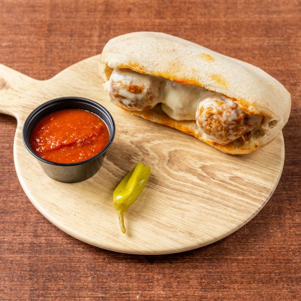 Meatball sub with melted cheese, a side of marinara sauce, and a pepper on a wooden board.