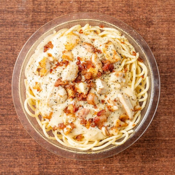 Creamy spaghetti carbonara with chicken and bacon bits, topped with herbs, in a clear bowl.
