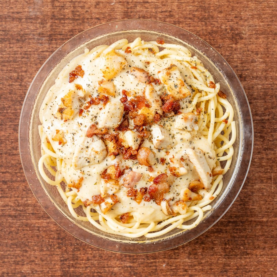 Creamy spaghetti carbonara with chicken and bacon bits, topped with herbs, in a clear bowl.