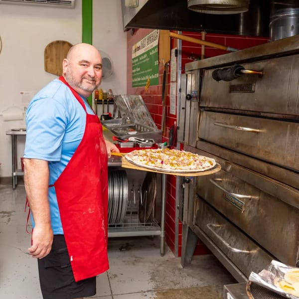 Grampie's Pizzeria chef in a red apron holding a raw pizza on a peel, ready for the oven.